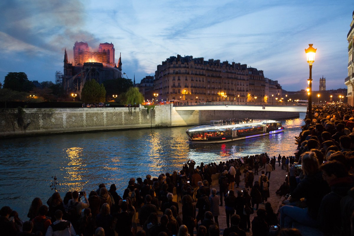 Bystanders look on as flames and smoke are seen billowing from the roof at Notre-Dame Cathedral with the Seine and boats by sun set river in Paris on April 15, 2019.